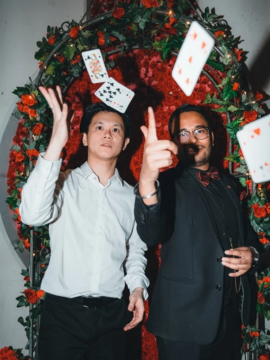 Two men standing in front of a heart-shaped backdrop decorated with red flowers, playing with floating playing cards.