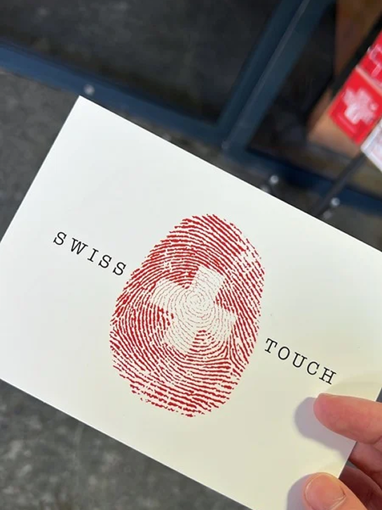 A white card with a red fingerprint featuring a white cross in the center. The words 'SWISS TOUCH' are printed on either side of the fingerprint.