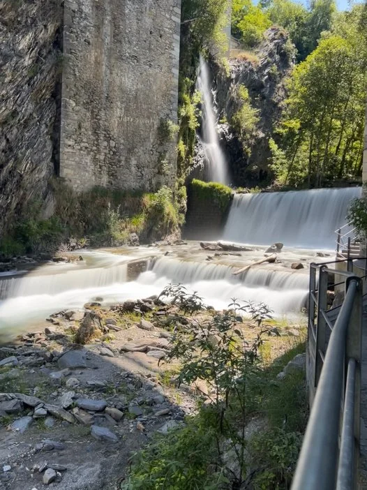 A flowing waterfall and river next to a stone wall and a pathway with a railing, surrounded by green trees and foliage.