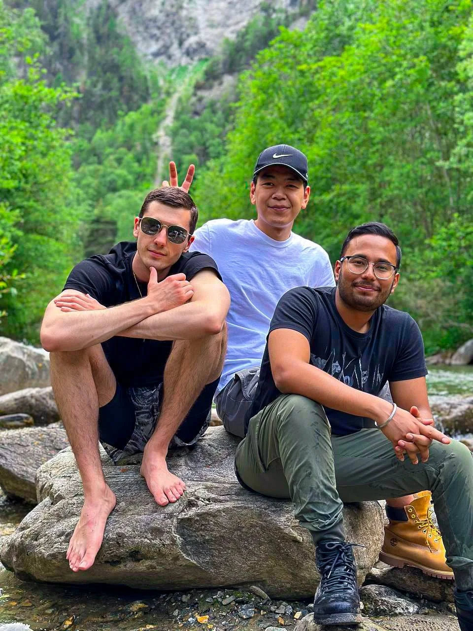Three men sitting on rocks by a river in a forested mountain area. One man is crouching with his arms crossed, wearing sunglasses and a black t-shirt. The second man, standing behind him, is smiling, wearing a white t-shirt and a black Nike cap, with bunny ears playful gesture behind the first man's head. The third man, sitting on the rock, has glasses, a dark t-shirt, and green pants, smiling with his hands clasped.