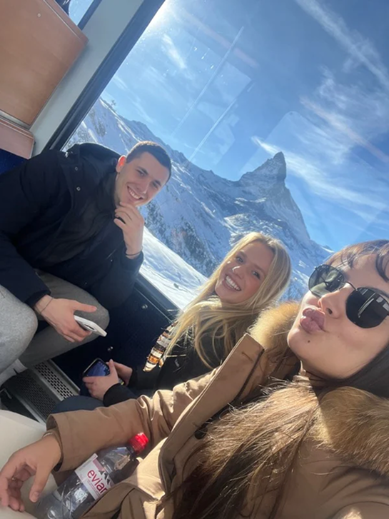 Three friends taking a selfie on a cable car with a snowy mountain and blue sky in the background.
