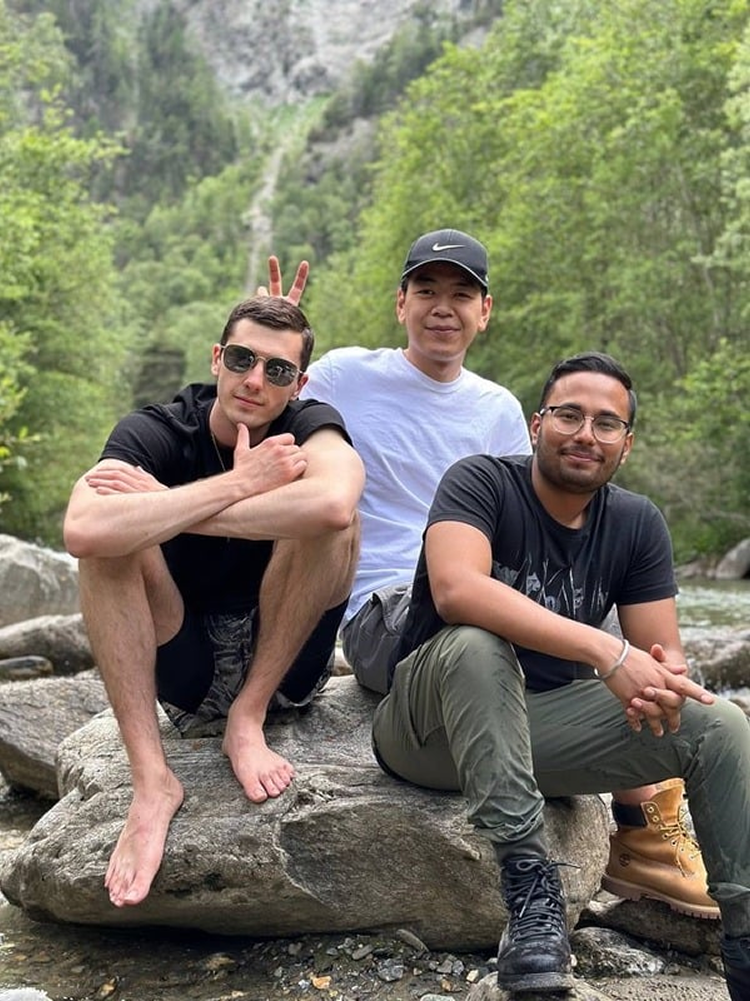 Three young men sitting on a large rock by a river in a forested area. One man is barefoot, wearing sunglasses, and making a peace sign behind another man with a cap. The third man is wearing glasses, sitting cross-legged, and smiling.