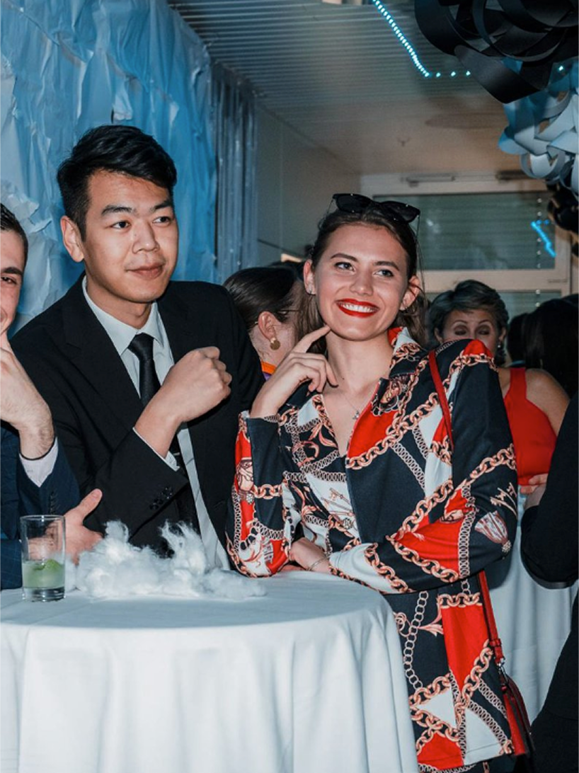 A group of people at a social event, with a young woman smiling and pointing to her face, wearing a patterned dress. A young man in a suit is standing next to her, and others are in the background. There is a table with a drink and some fog effects on it.