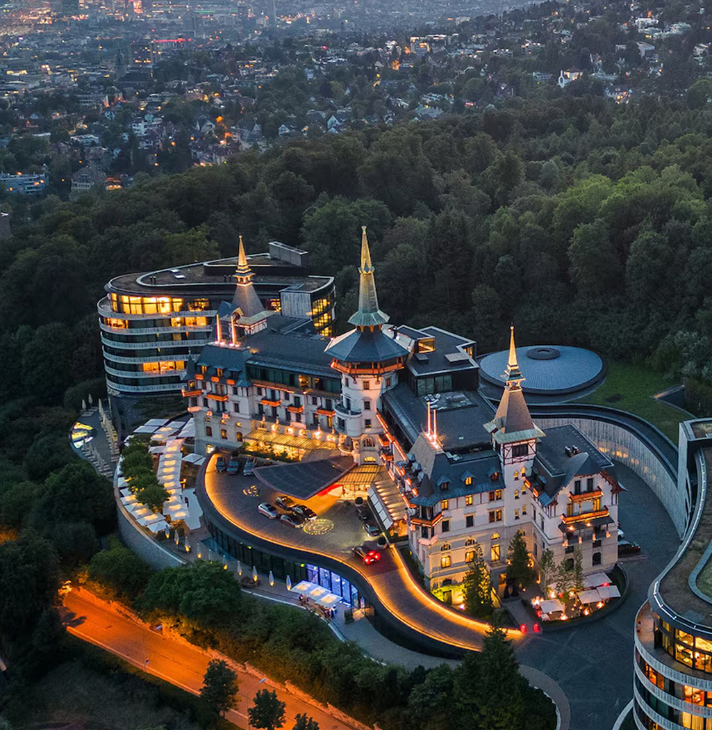An elegant fantasy castle hotel with turrets and spires, illuminated at dusk, surrounded by lush greenery and a cityscape in the distance.