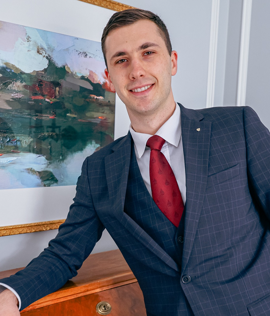 A young man in a dark blue checkered suit, white shirt, and red tie, smiling and leaning on a wooden table in an office. There is an abstract painting in the background.