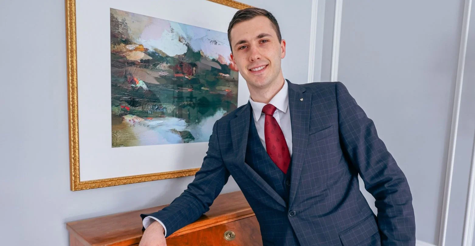 A young man in a blue checkered suit with a red tie smiling and leaning on a wooden surface indoors, with a colorful abstract painting on the wall behind him.