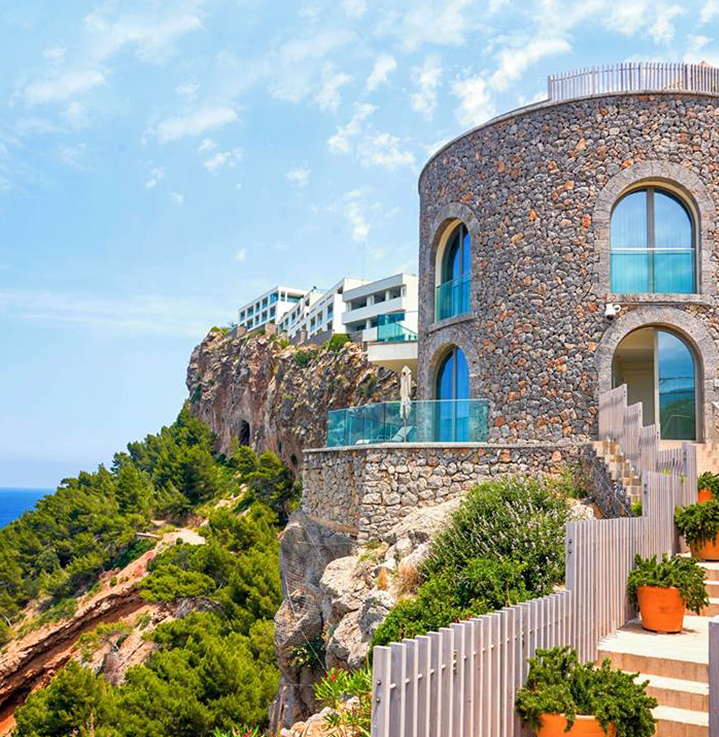 A stone house built into a cliffside with large arched windows, glass balconies, and a stairway leading down to a garden with potted plants, overlooking a lush green hillside and the ocean under a partly cloudy sky.
