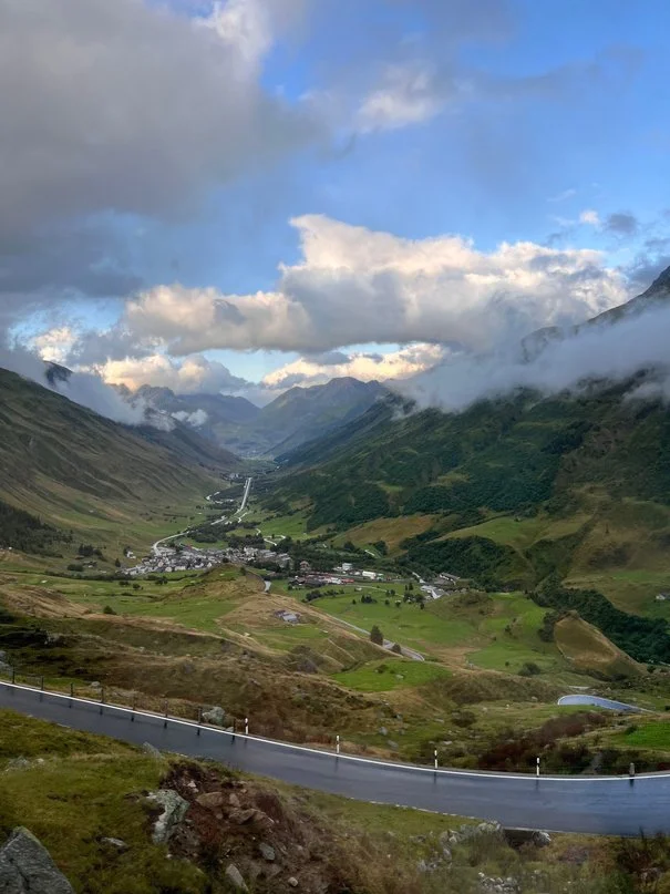 A scenic view of a mountainous valley with a winding road, green hills, scattered houses, and cloudy sky.