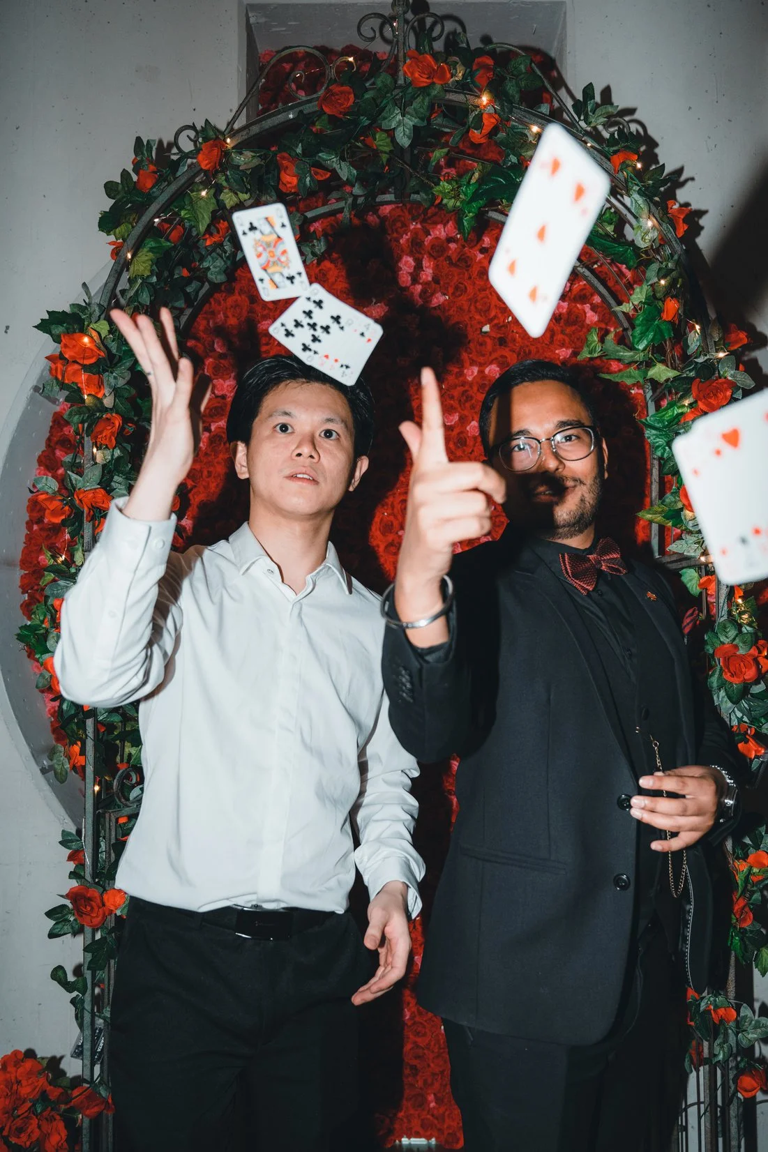 Two men dressed in formal attire standing in front of a decorative red floral wall, with playing cards flying in the air around them. One of the men is wearing a white shirt and the other a black suit with a bow tie. The man in black is pointing at the camera.