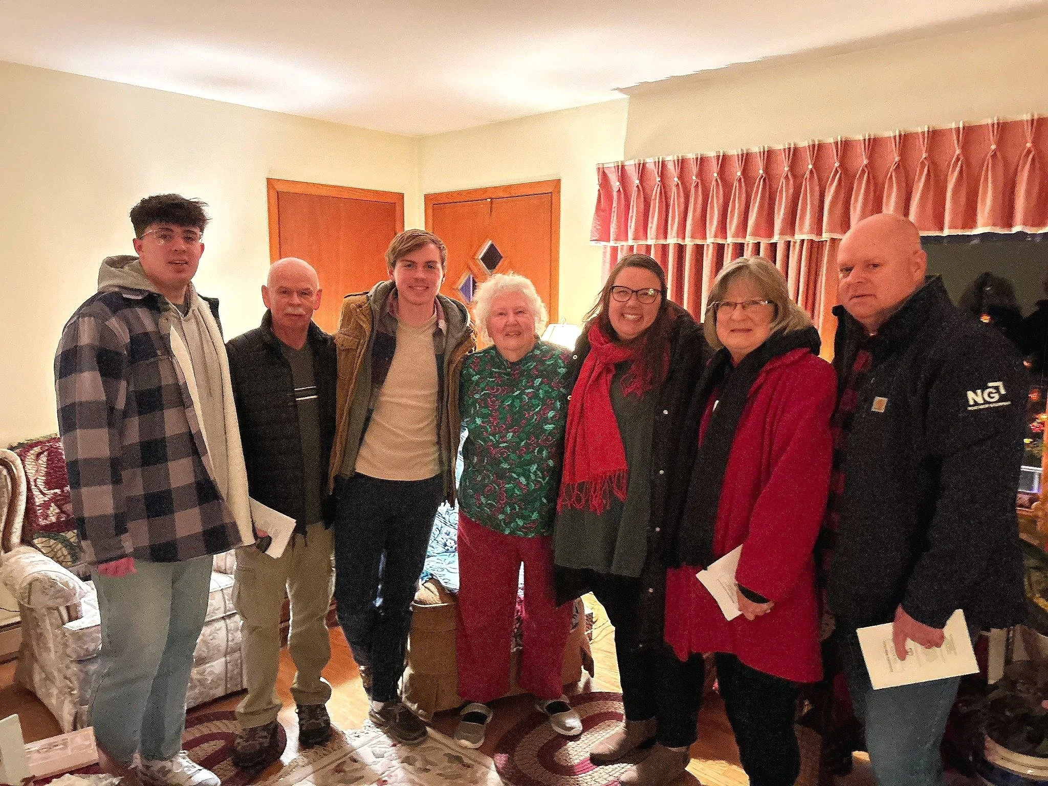 Group of seven people standing together in a living room, smiling for the camera. The group includes young adults, middle-aged, and elderly, with a mix of men and women. The room has beige walls, a floral-patterned sofa, and red curtains.