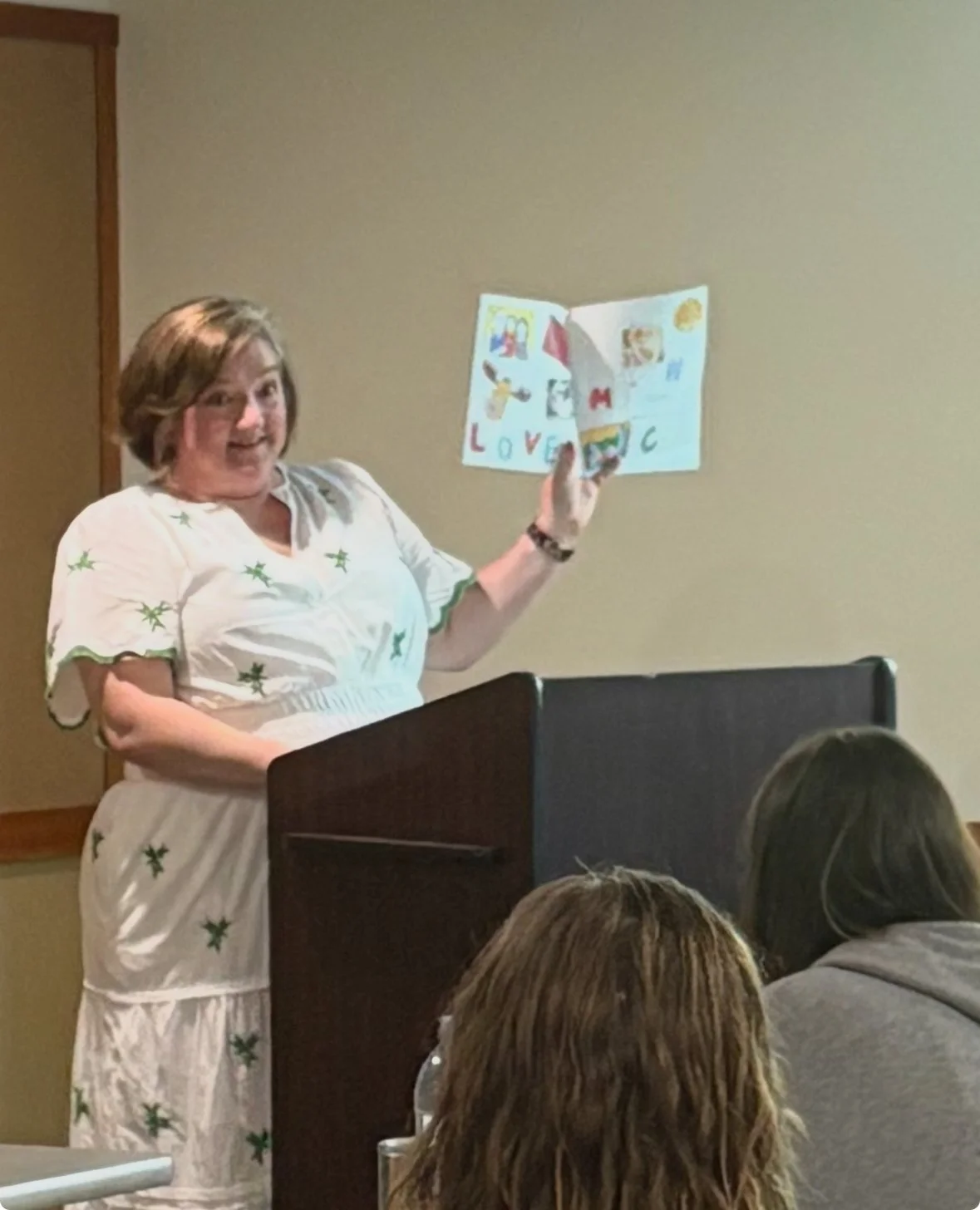 A woman in a white dress with green star patterns is standing at a podium, holding up a colorful children's drawing and smiling at an audience.