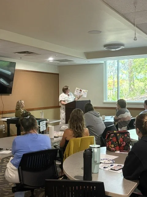 A woman is giving a presentation in a conference room with a large window that overlooks trees outside. Attendees are seated at tables, listening.