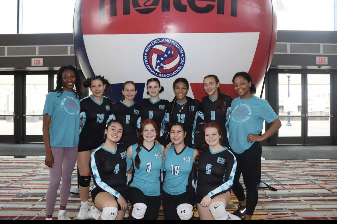 Group of young female volleyball players and coaches posing for a team photo indoors, with a large volleyball graphic and a Northeast Volleyball Association logo in the background.