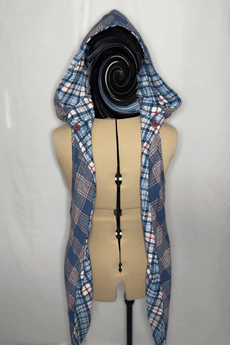 A dress form with a black swirl for a head, dressed in a plaid shirt with a hood and a black strap in the center.