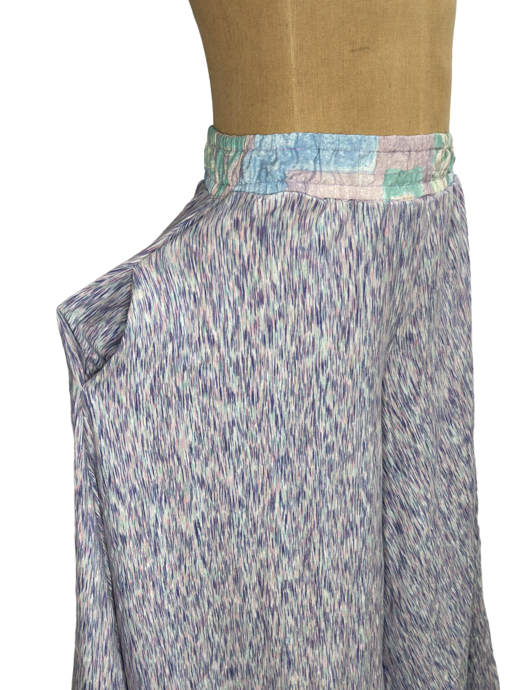 Side view of palazzo pants in a purple space-dyed print with a pastel patchwork elastic waistband, teal hem trim, and an open side pocket, shown on a dress form against 