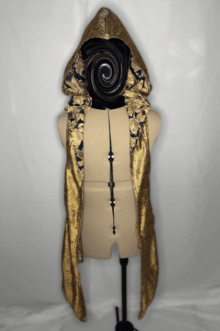 A mannequin wearing a gold and black patterned hooded textile with gold accents, draped over a beige torso form against a plain grey background.