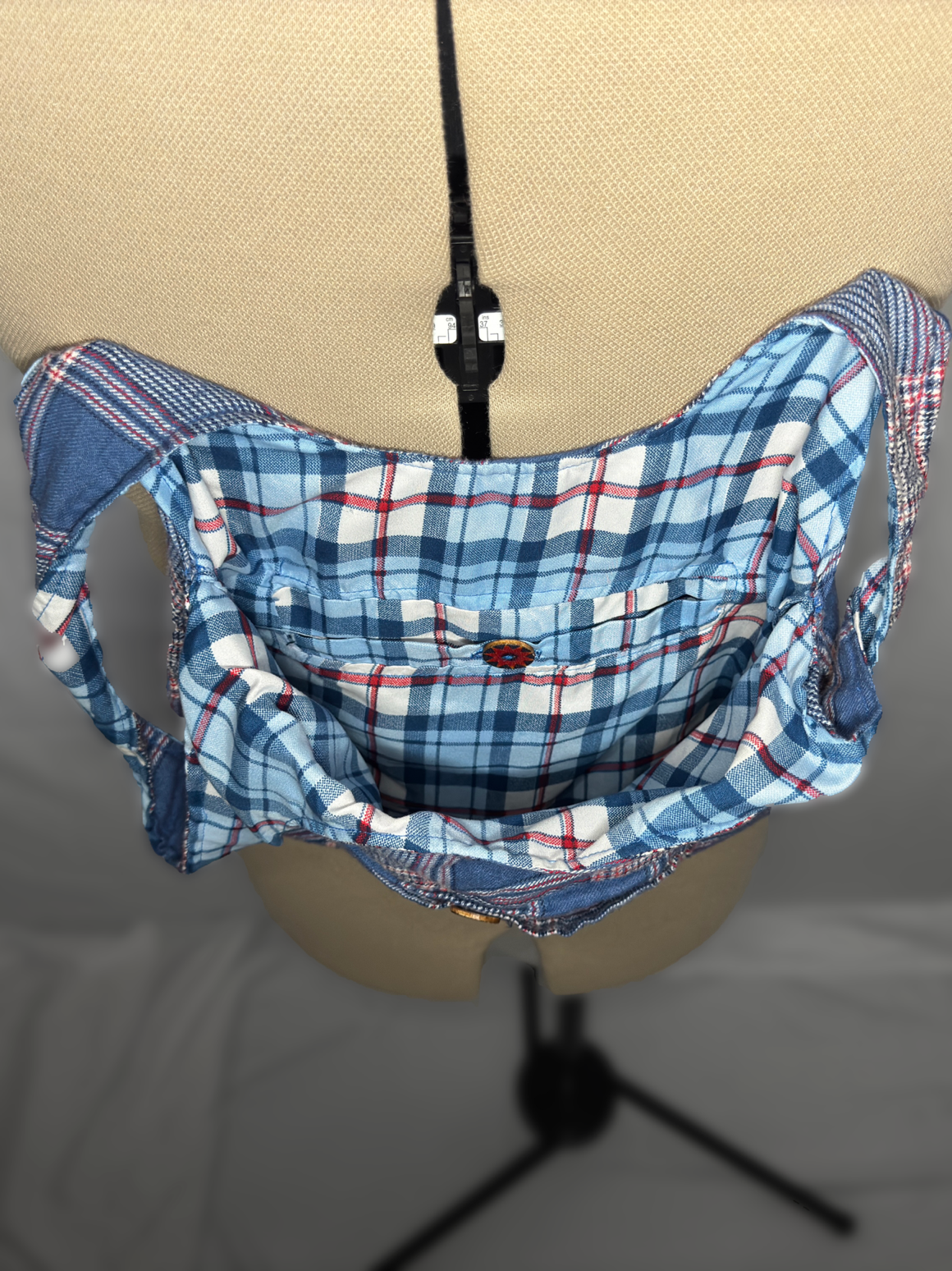 A beige mannequin torso on a black stand wearing a plaid shirt turned collarless, revealing a black zipper.