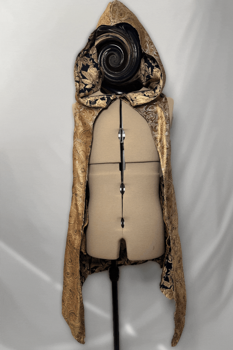 A mannequin with a decorative cloak hanging over its shoulders, a spiral design on the head, and a beige torso form on a stand against a plain background.