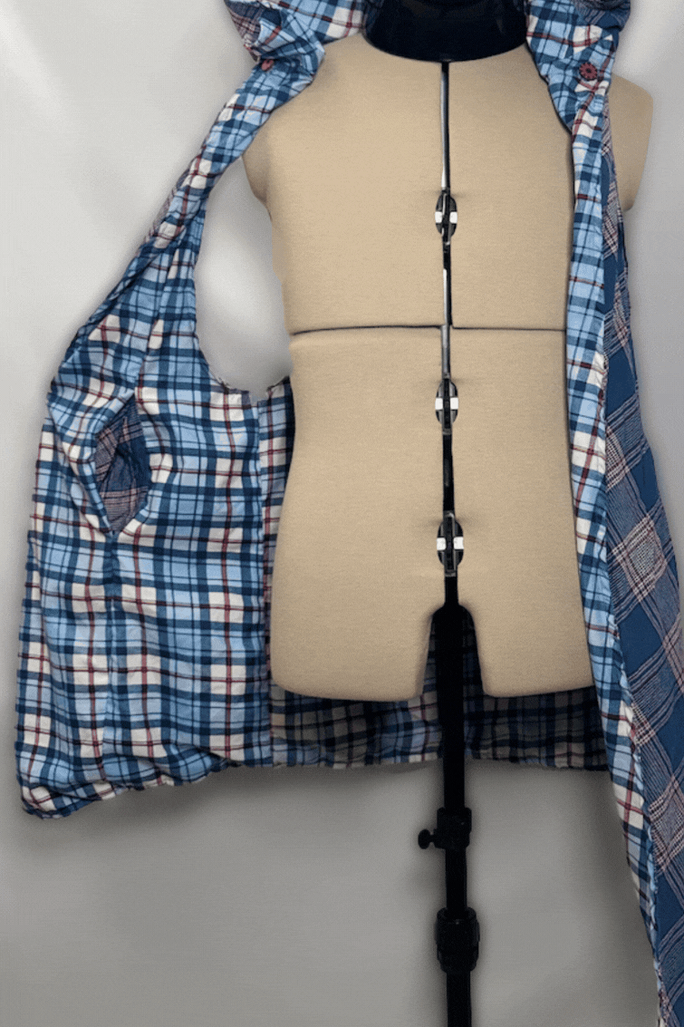 A mannequin torso on a stand, wearing a blue plaid shirt open at the front with a black zipper, and a beige padded section on the front for fitting clothing or accessories.