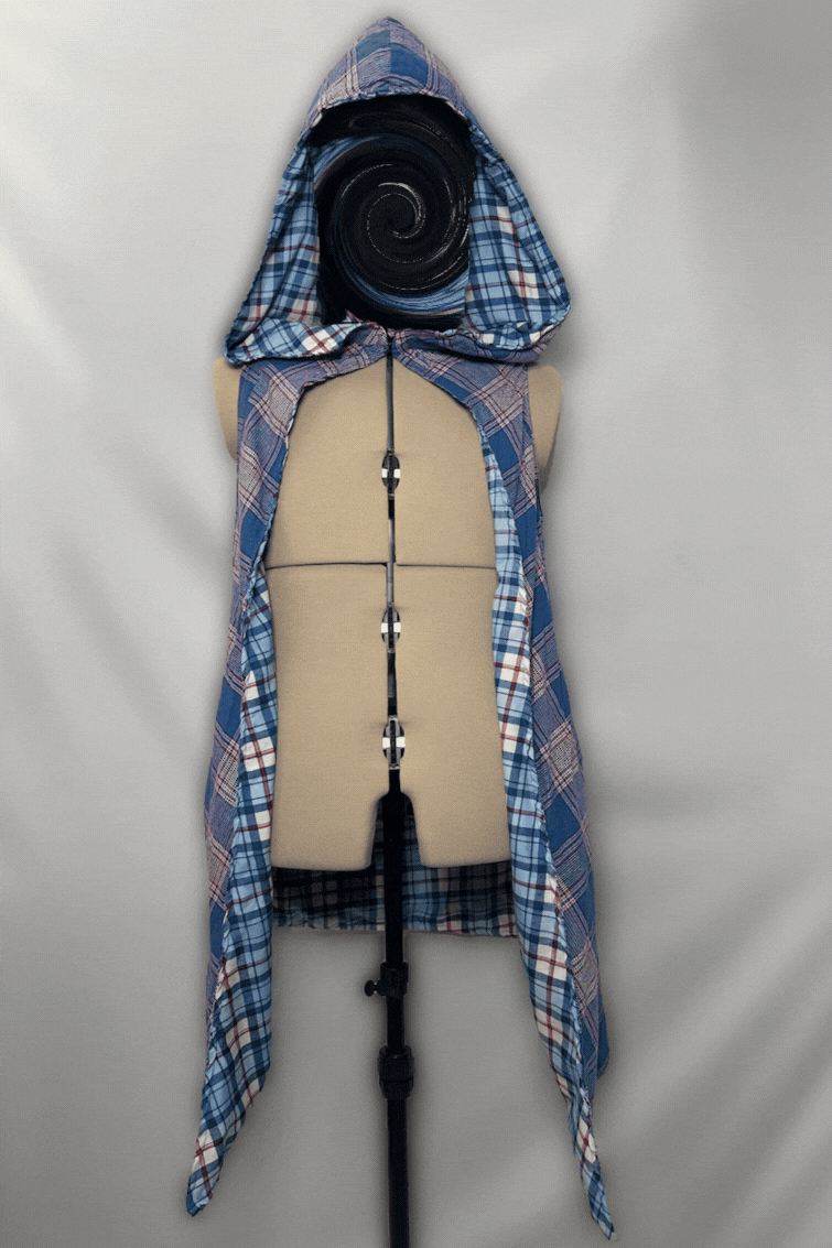 A dress form mannequin wearing a layered costume with a plaid shirt and a hooded cape or cloak, with a background of neutral fabric.