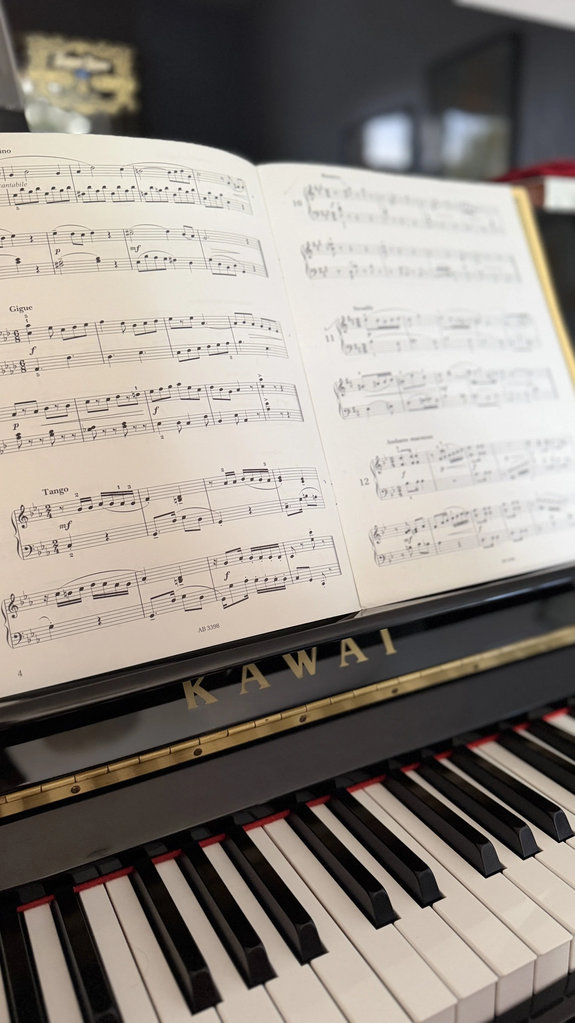 Close-up of a Kawai upright piano with an open sheet music book on top, displaying musical notes and staff lines. The piano's keys are visible at the bottom of the image. The book is a snapshot of ABRSM Sight Reading exercises.