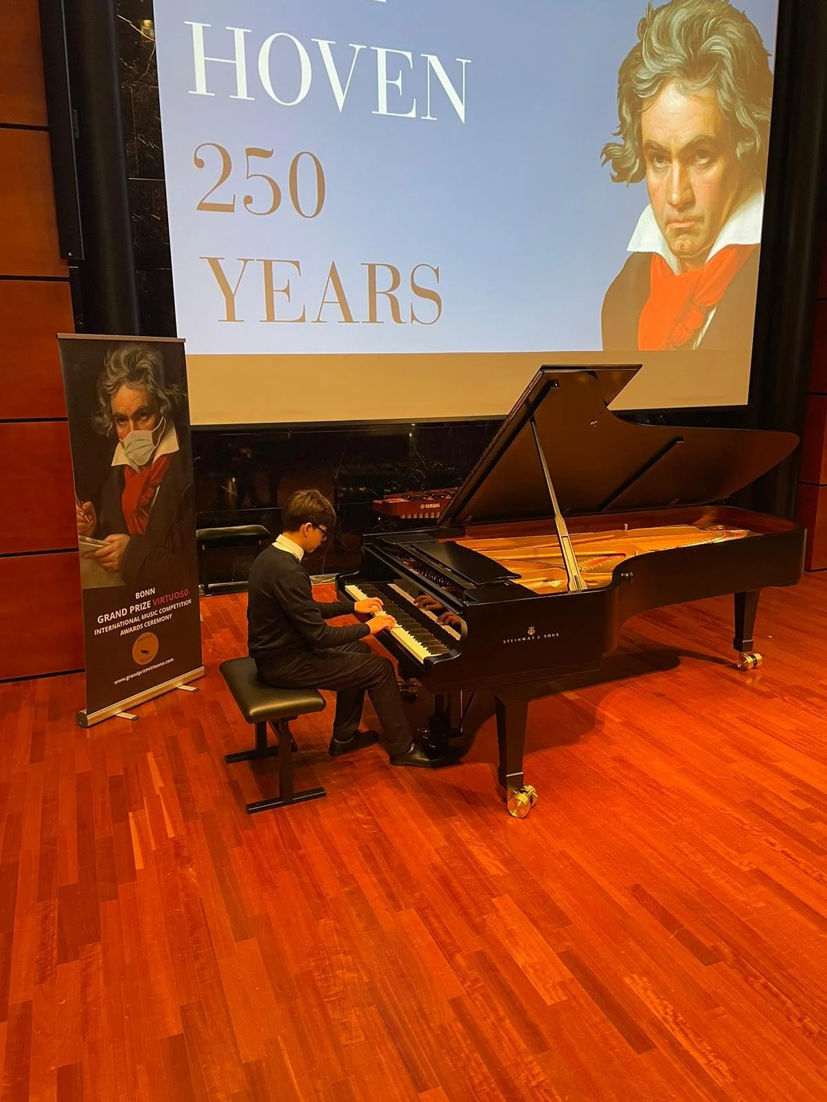 A young man playing a grand piano on a stage with a large screen behind him that displays a picture of Beethoven. There is a vertical banner to his left with Beethoven's image wearing a face mask and text about a music competition award ceremony.