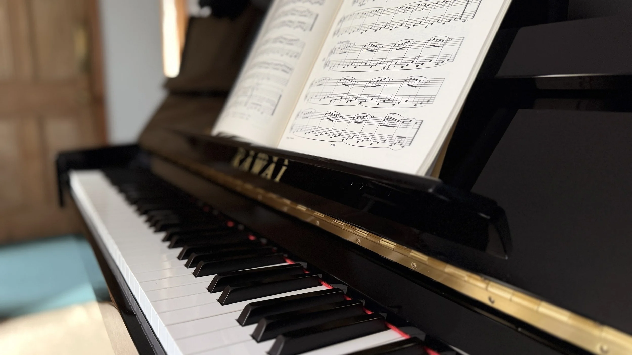 Close-up of a black Kawai upright piano with Beyer's Preparatory School music book with an open bright corridor in the background.