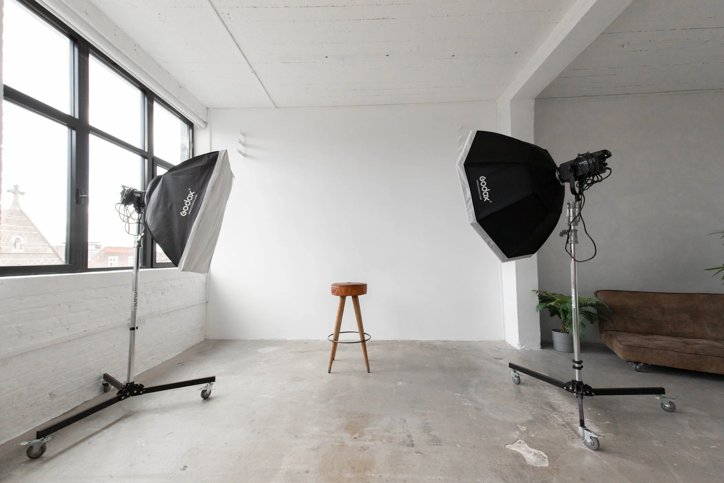 Photography studio with two softbox lights on stands and a brown stool in front of a white wall, with a window and a plant on the right side.