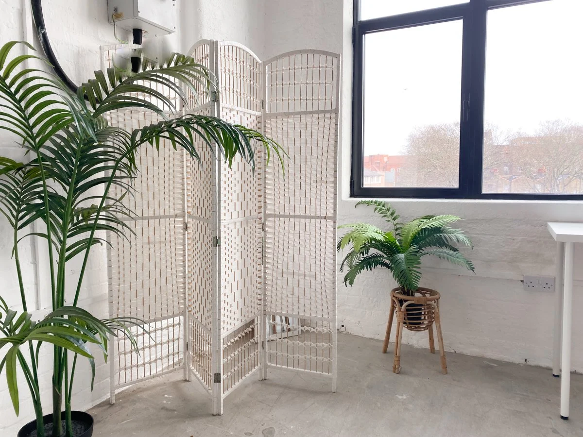 Interior space with large potted plants, a wooden decorative screen divider, a window with a view of trees and buildings outside, white painted brick wall, concrete floor, and a white table leg.