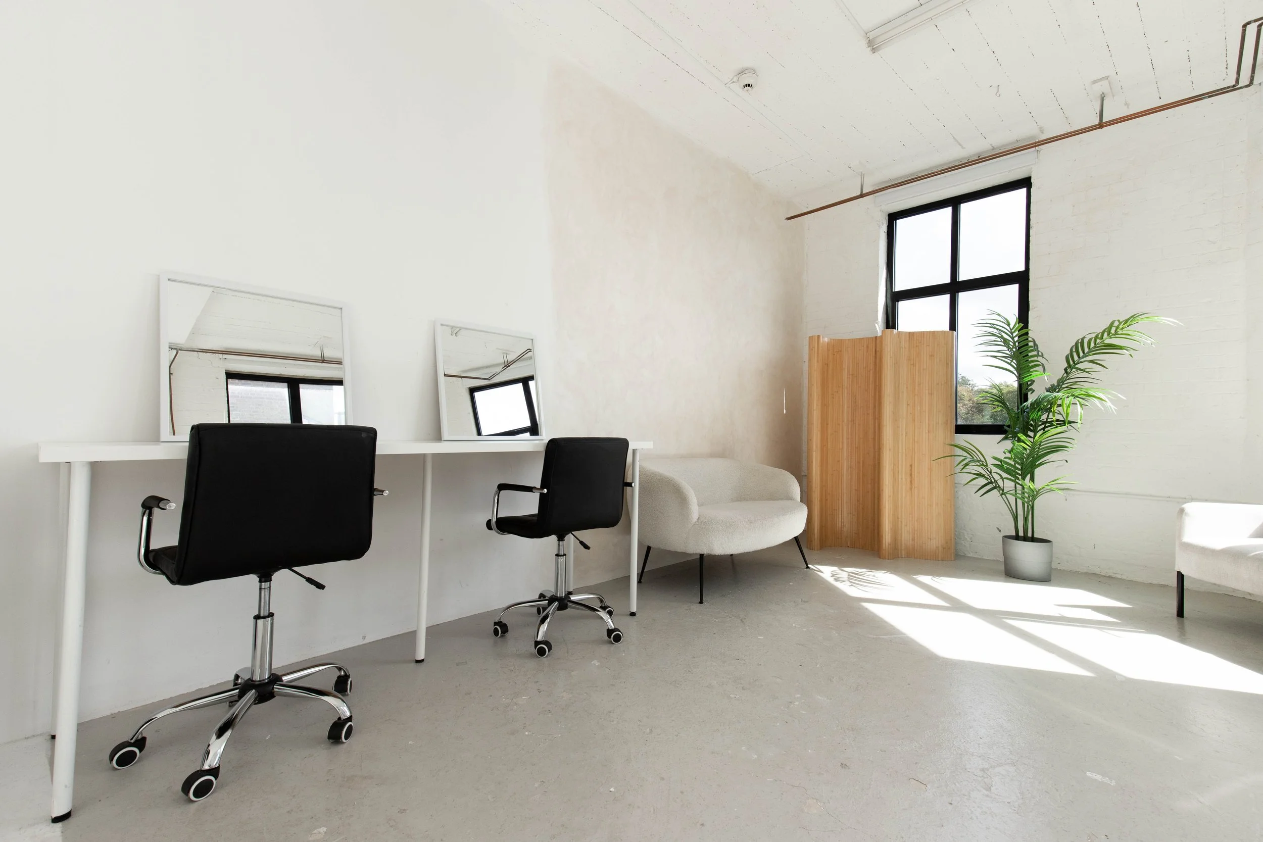 Minimalist office space with two black swivel chairs, white desk, two mirrors, a beige sofa, a wooden room divider, a potted plant, a large window, and a white brick wall.