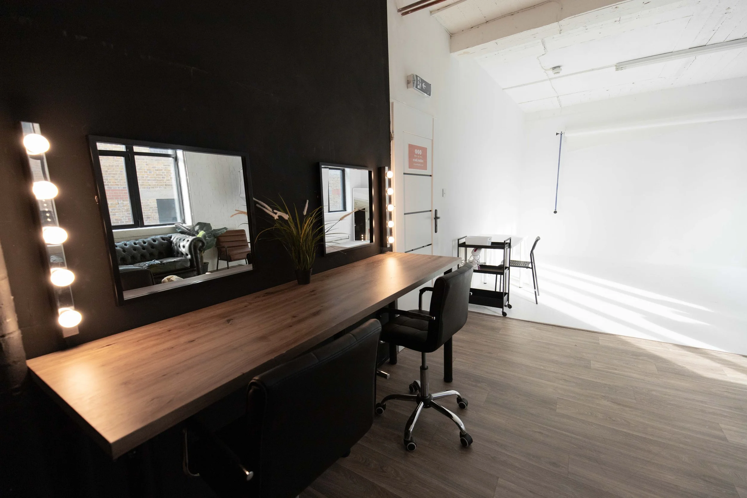 A room with a black accent wall featuring two mirrors, a wooden table with a potted plant, black chairs, and a vanity mirror with lights. The space has large windows reflecting an outdoor building.