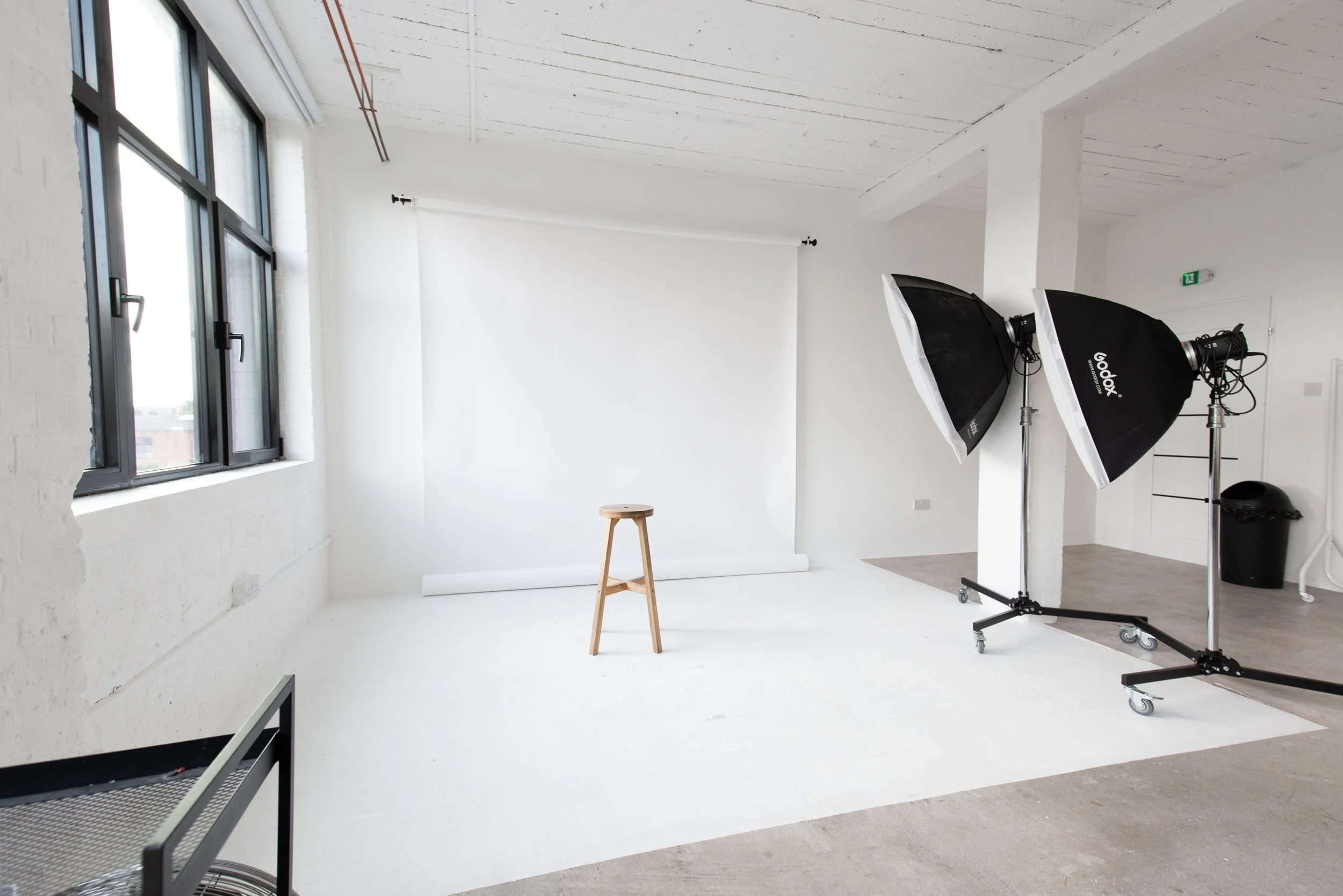 Photography studio with white backdrop, two softbox lights, a wooden stool, large window with black frame, and a trash bin.