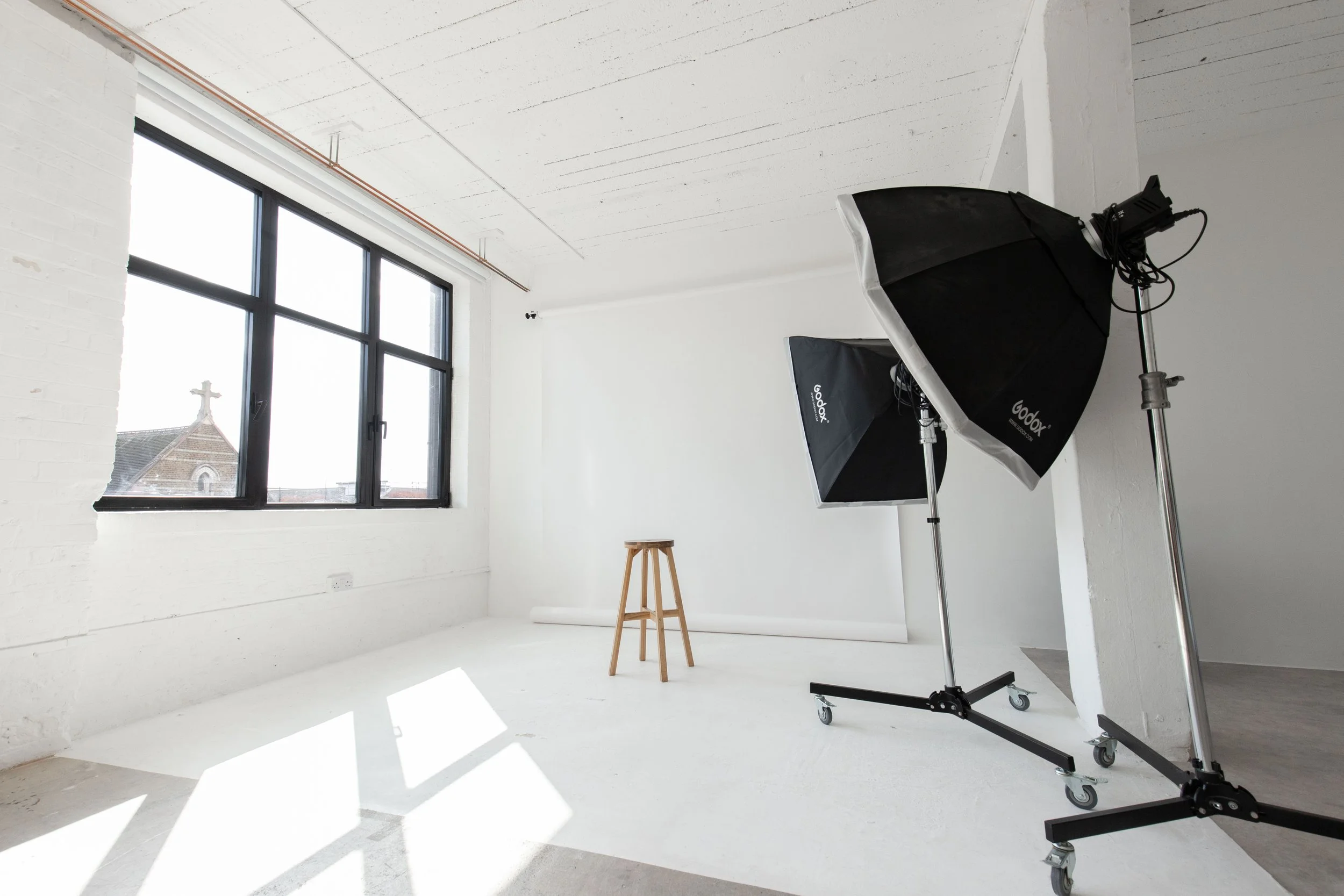 Photography studio with white walls, large window letting in natural light, two studio lights with umbrellas, a wooden stool, and a white backdrop.