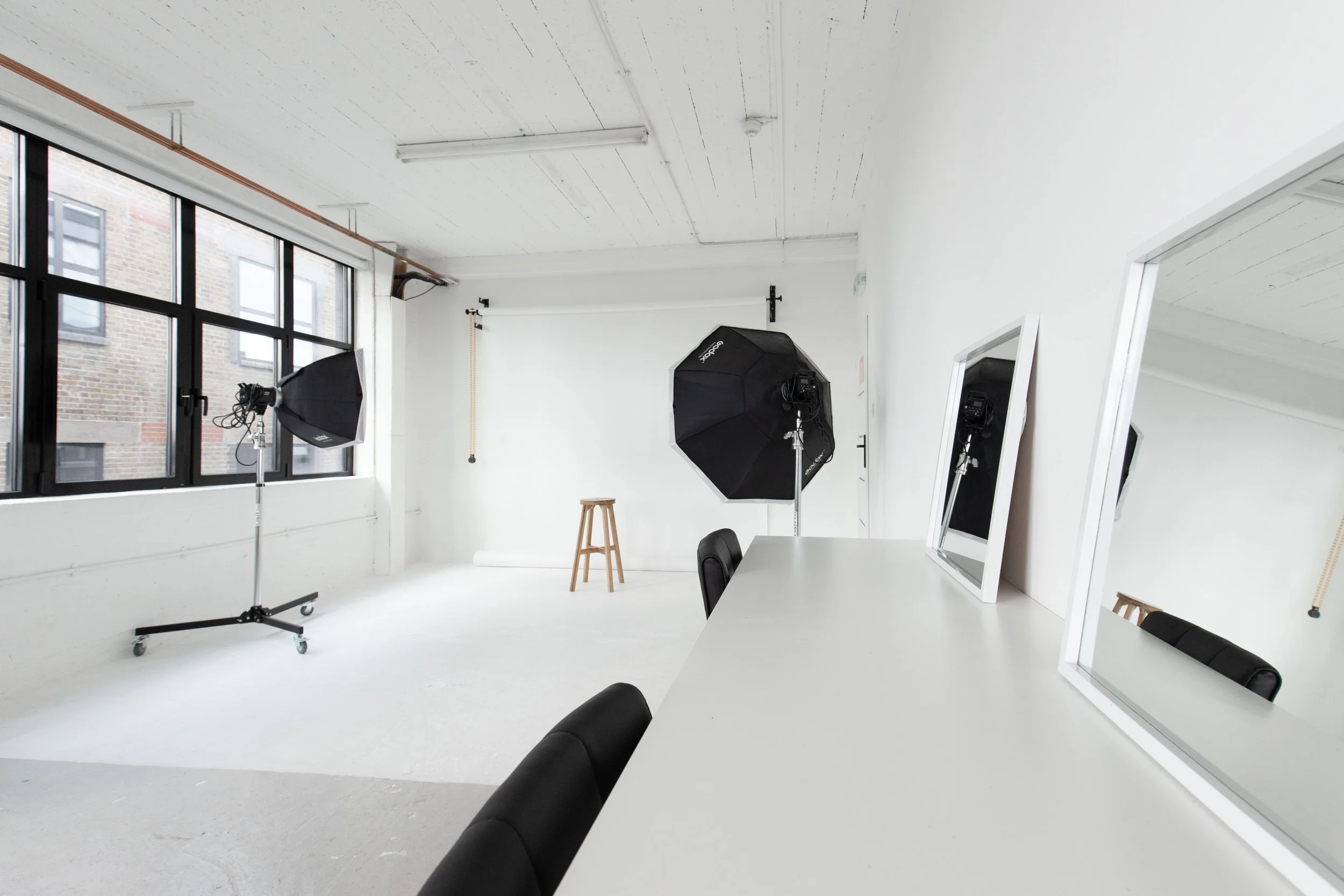 Photography studio with white walls and ceiling, large window with black frames, two softbox lights, a stool, a white table with black chairs, and two mirrors, one leaning against the table and the other on the wall.