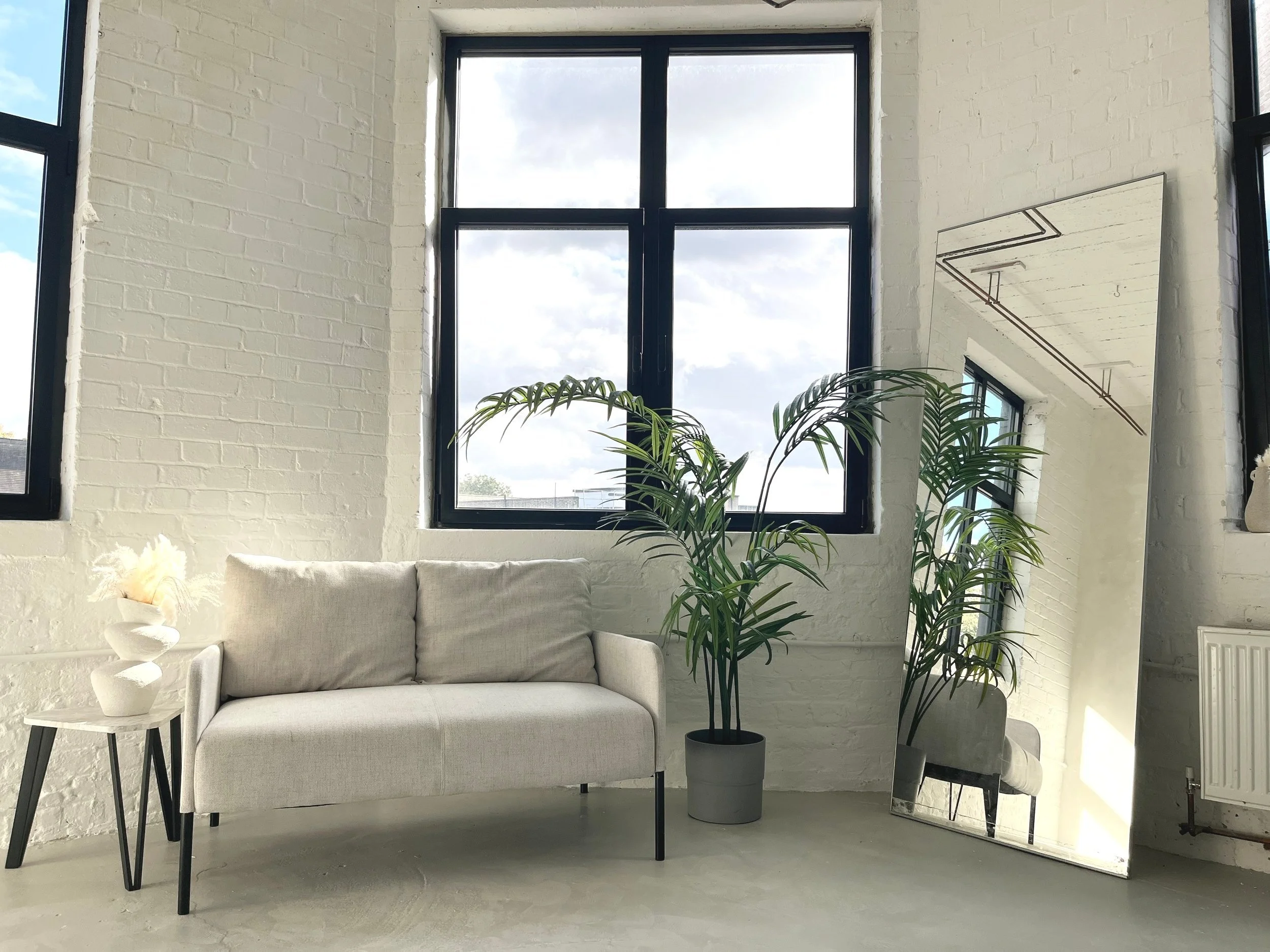 A white couch with black legs in front of a white brick wall, next to a potted plant and a tall mirror reflecting part of the room and window.