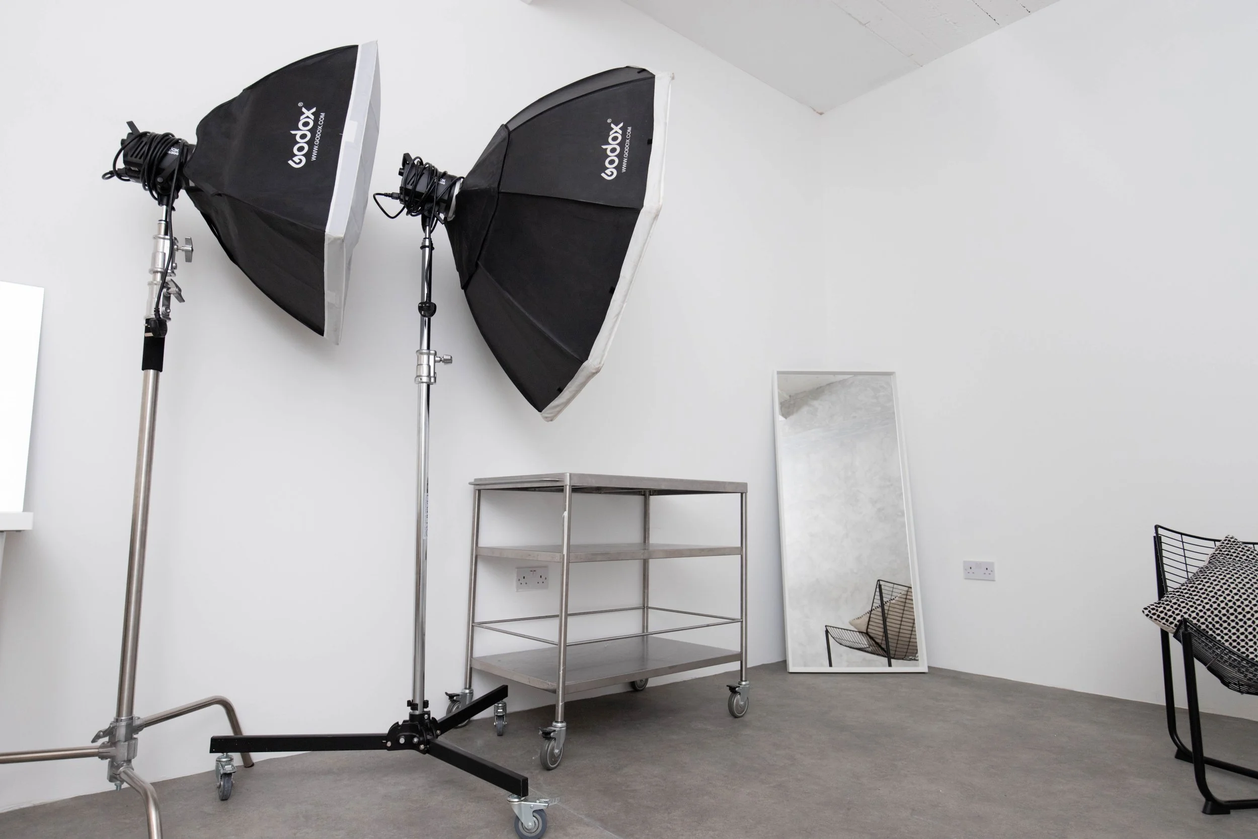 Photography studio space with two large Godox softbox lights on stands, a metal utility cart, a tall mirror leaning against the wall, and a black wire chair with a cushion, all in a room with white walls and grey carpet.