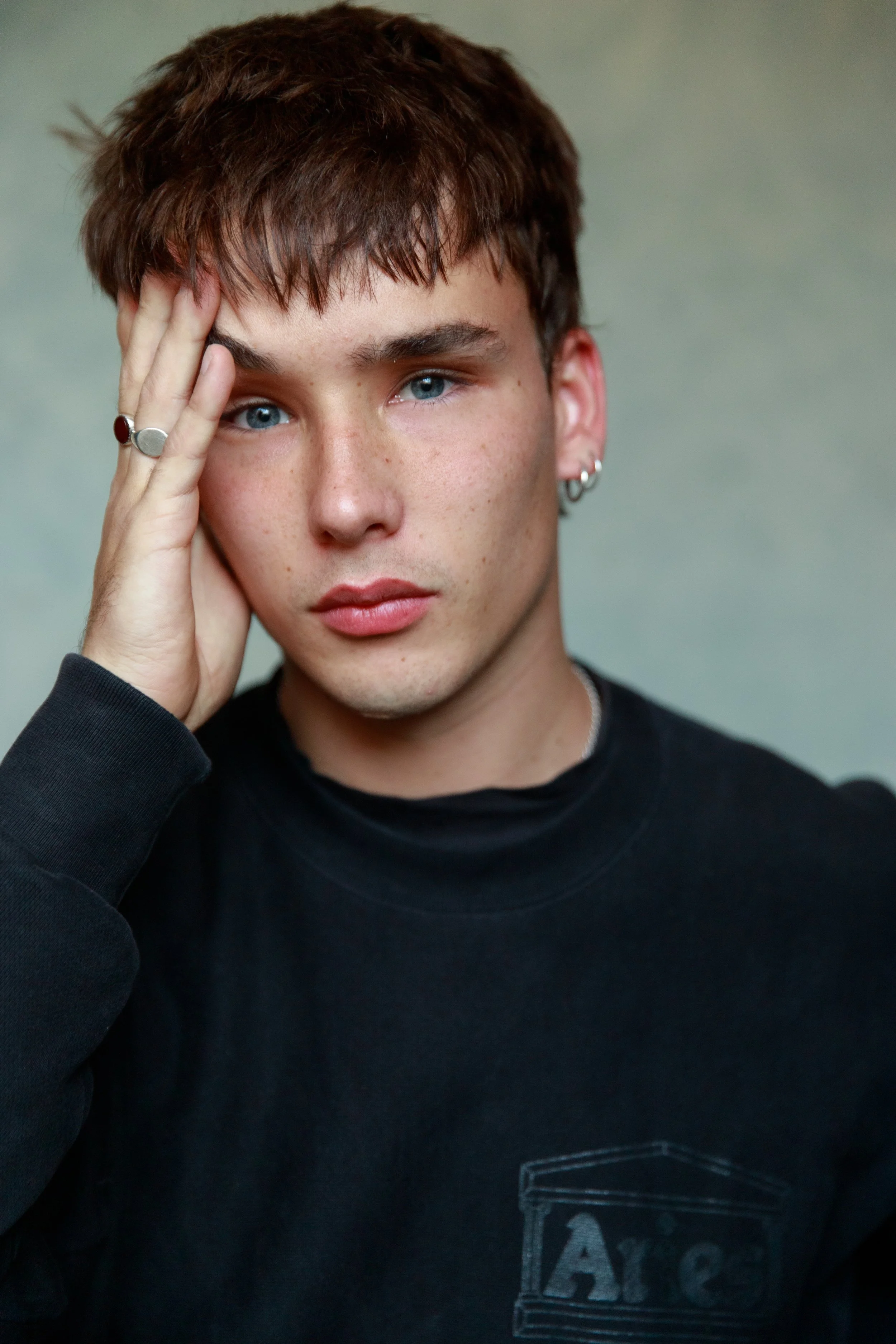 A young man with light skin and blue eyes, touching his forehead with his left hand, wearing a black sweatshirt with a logo, and earrings.