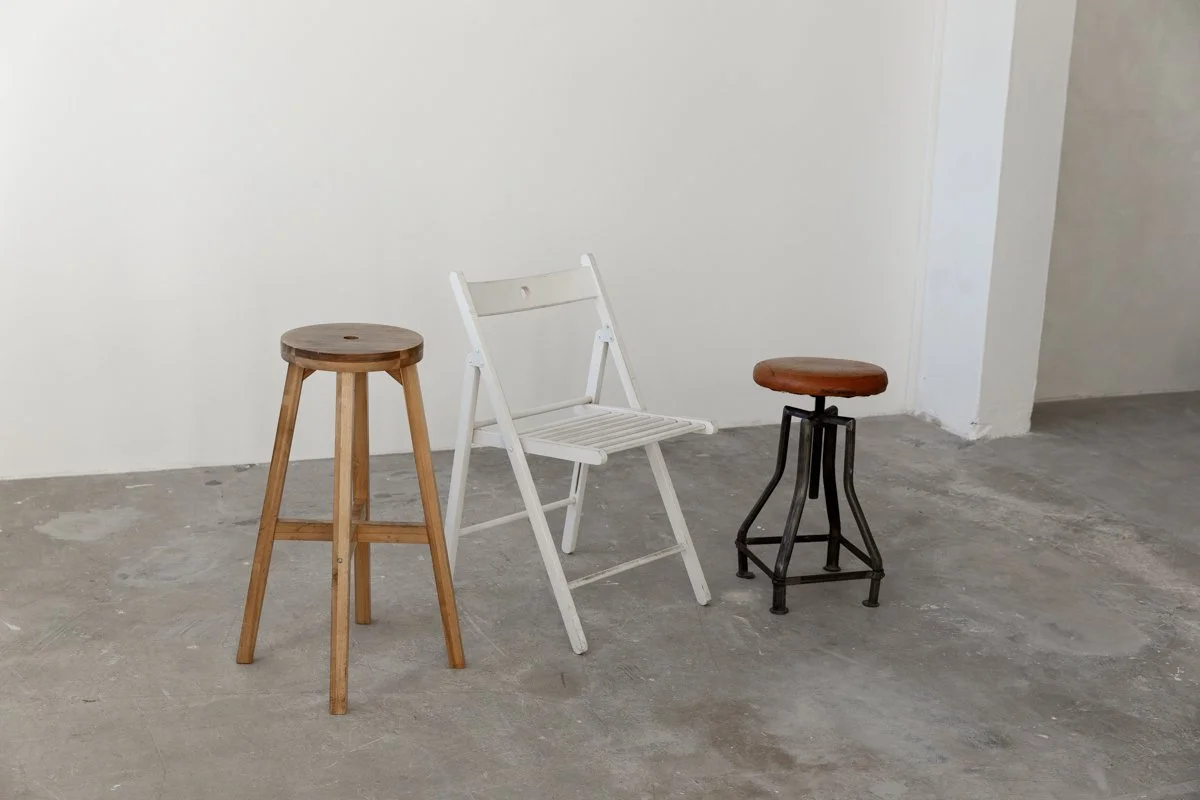 Three different style stools and a white folding chair placed on a concrete floor against a white wall.