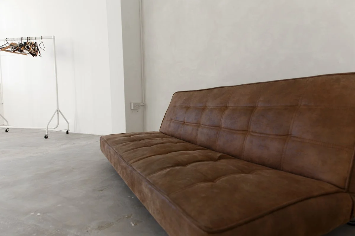 A brown leather sofa in a mostly empty room with a white wall and a clothing rack with hangers in the background.