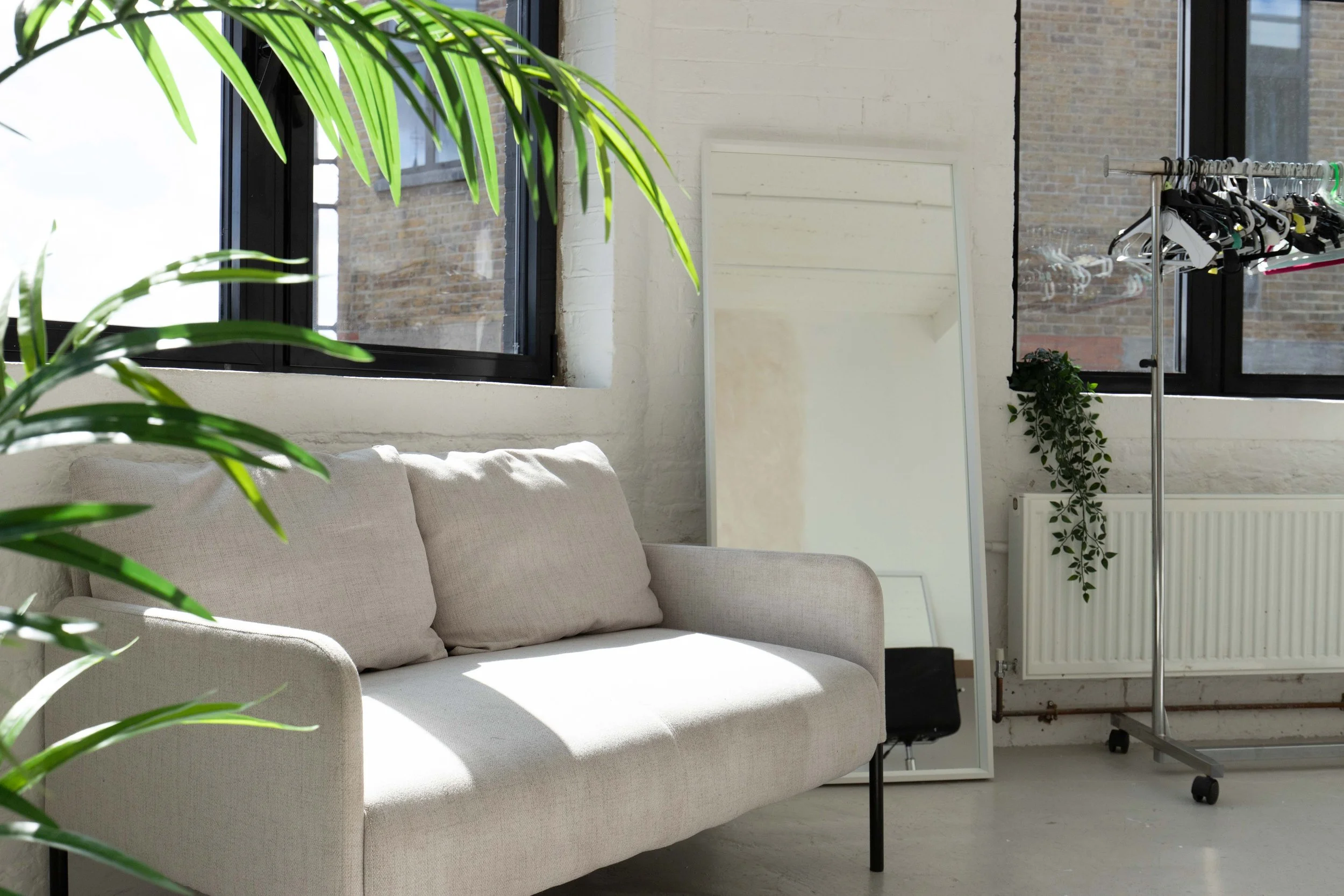 A bright, minimalistic room with a beige sofa, large windows, green plants, a full-length mirror, and a clothing rack.