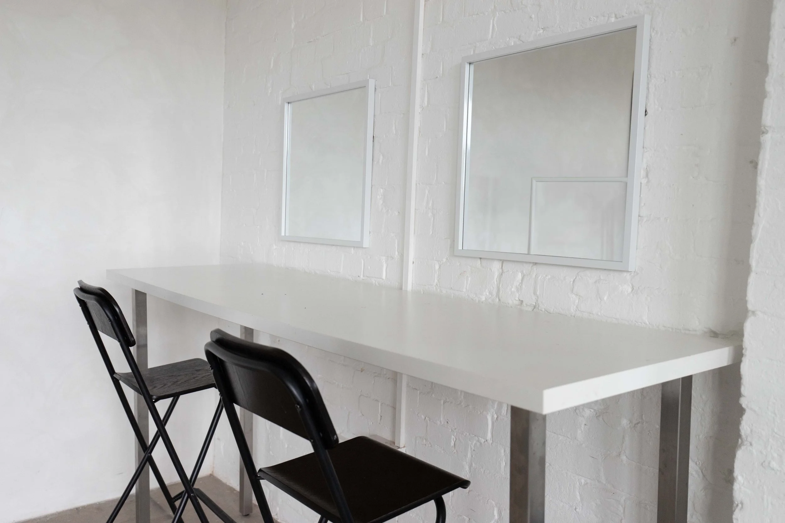 A white table with two black folding chairs in front, and two large white-framed mirrors on a white brick wall.