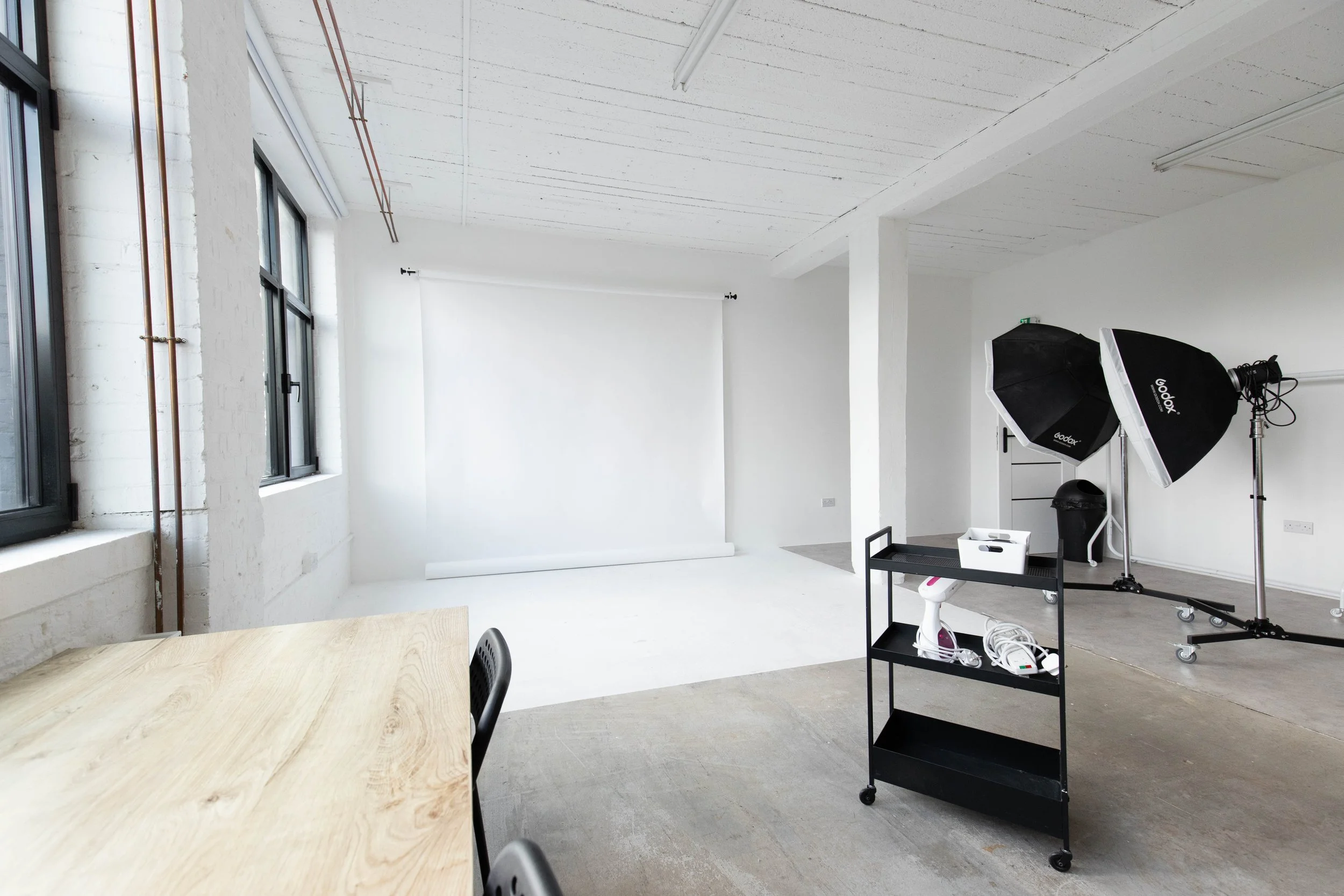 Photography studio setup with a white backdrop, large softbox lights, and a rolling cart with photography equipment in a bright industrial-style room.