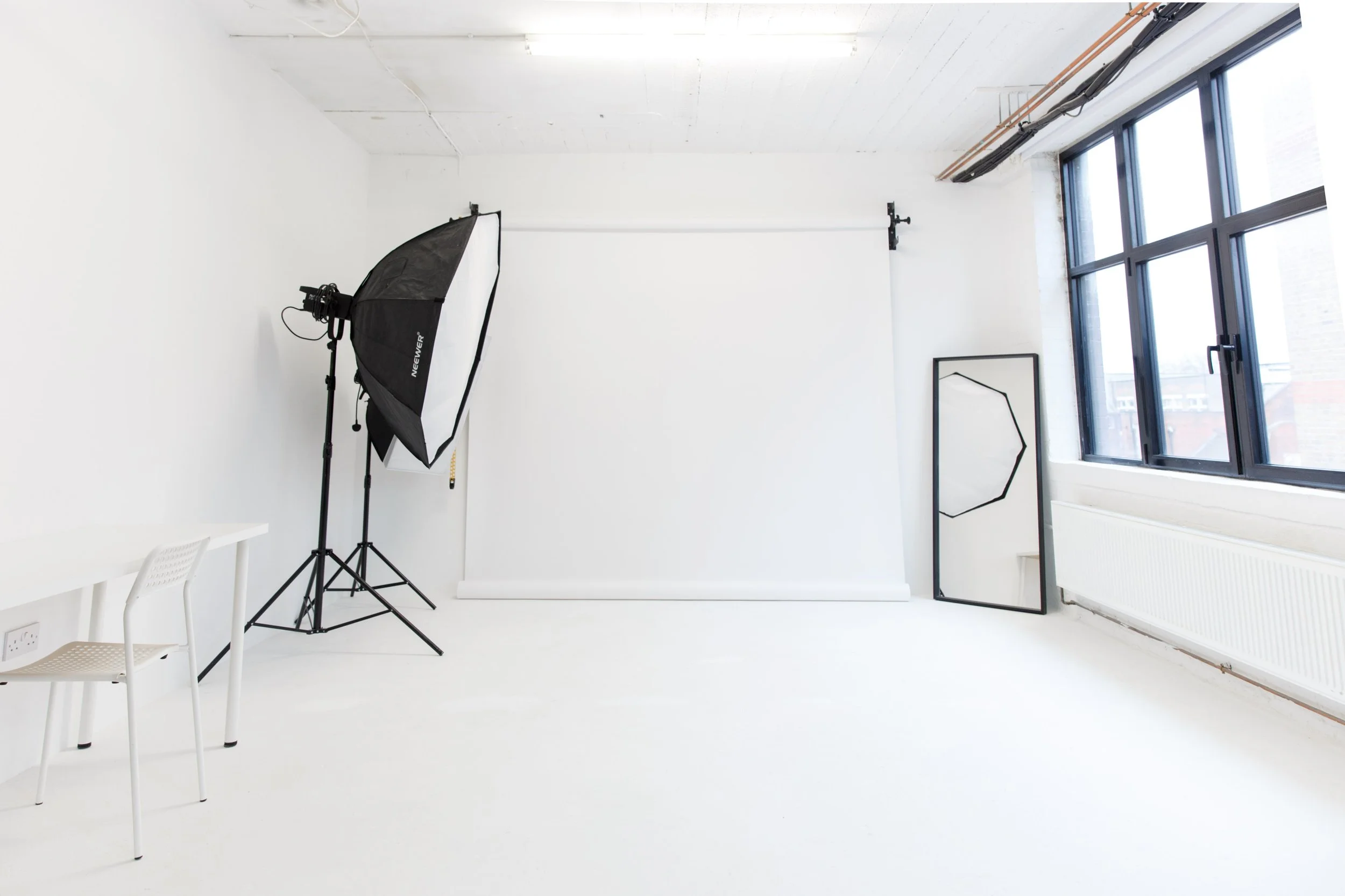 Photography studio with a white backdrop, large window with black frames, a standing mirror, and photography equipment including a softbox light