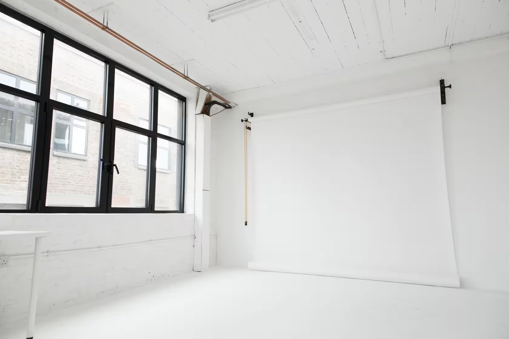 Empty photography studio with large black-framed windows, white walls, a white roll-up backdrop, and ceiling-mounted track for hanging backdrops.