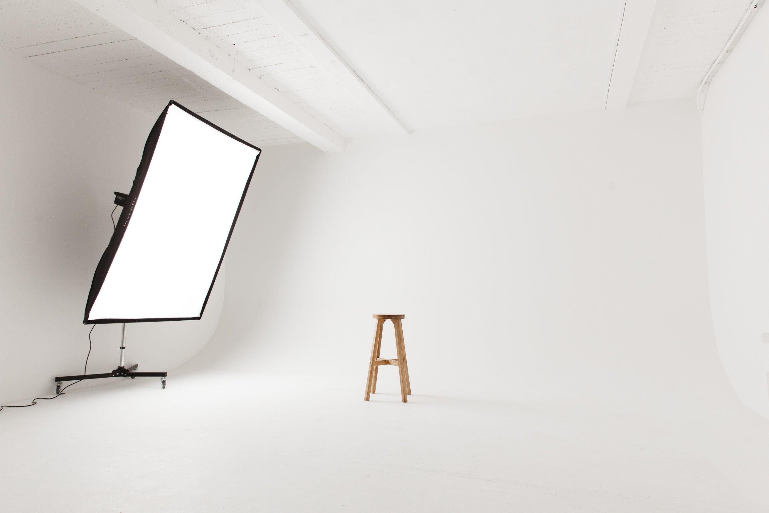 Photography studio with large softbox light and wooden stool on white backdrop.