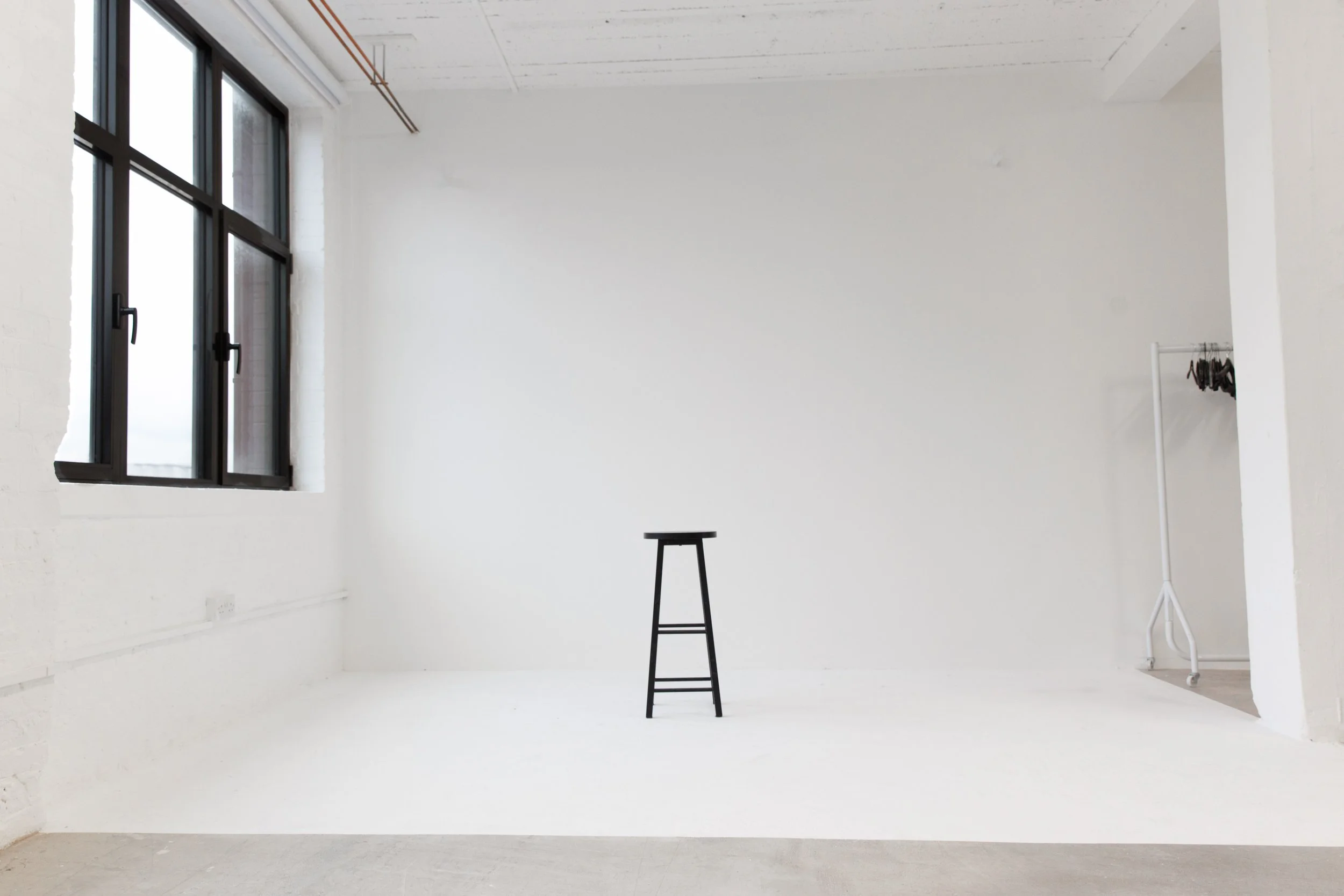 Interior of a mostly empty room with white walls, a large window on the left, a black stool in the center, and a clothing rack on the right.