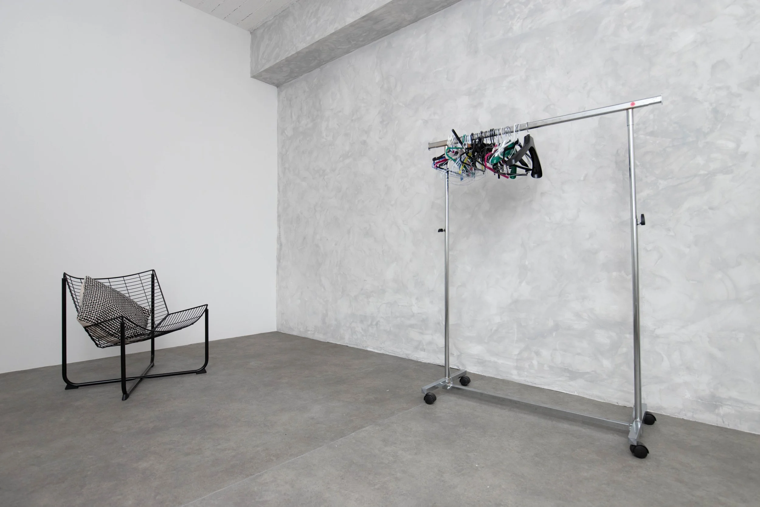 Minimalist room with a black wire chair and a metal clothing rack holding hangers, situated against textured white and gray walls with a concrete floor.