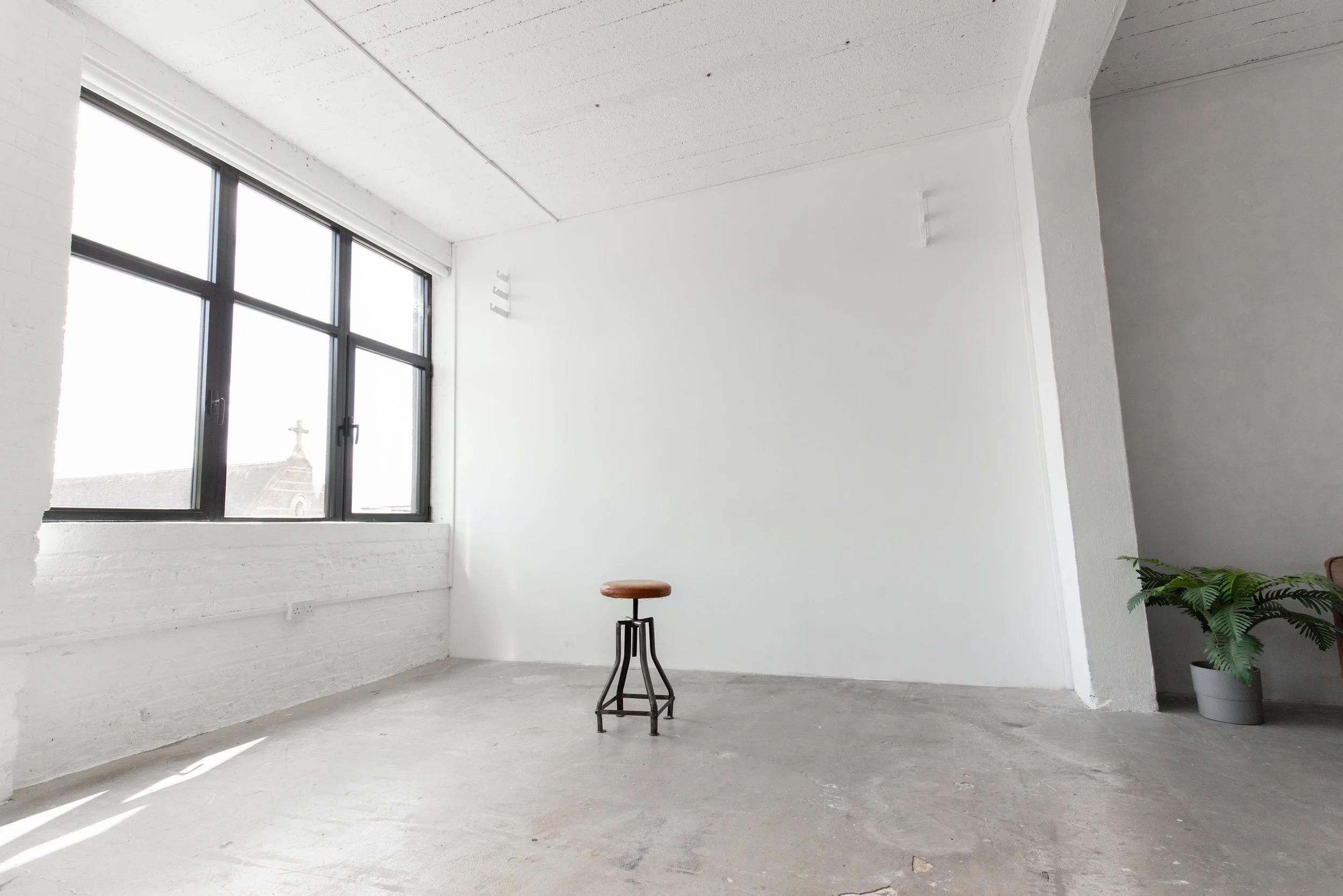 Empty room with large window, white brick wall, concrete floor, and a single stool near the center. A potted plant is in the corner near the right wall.