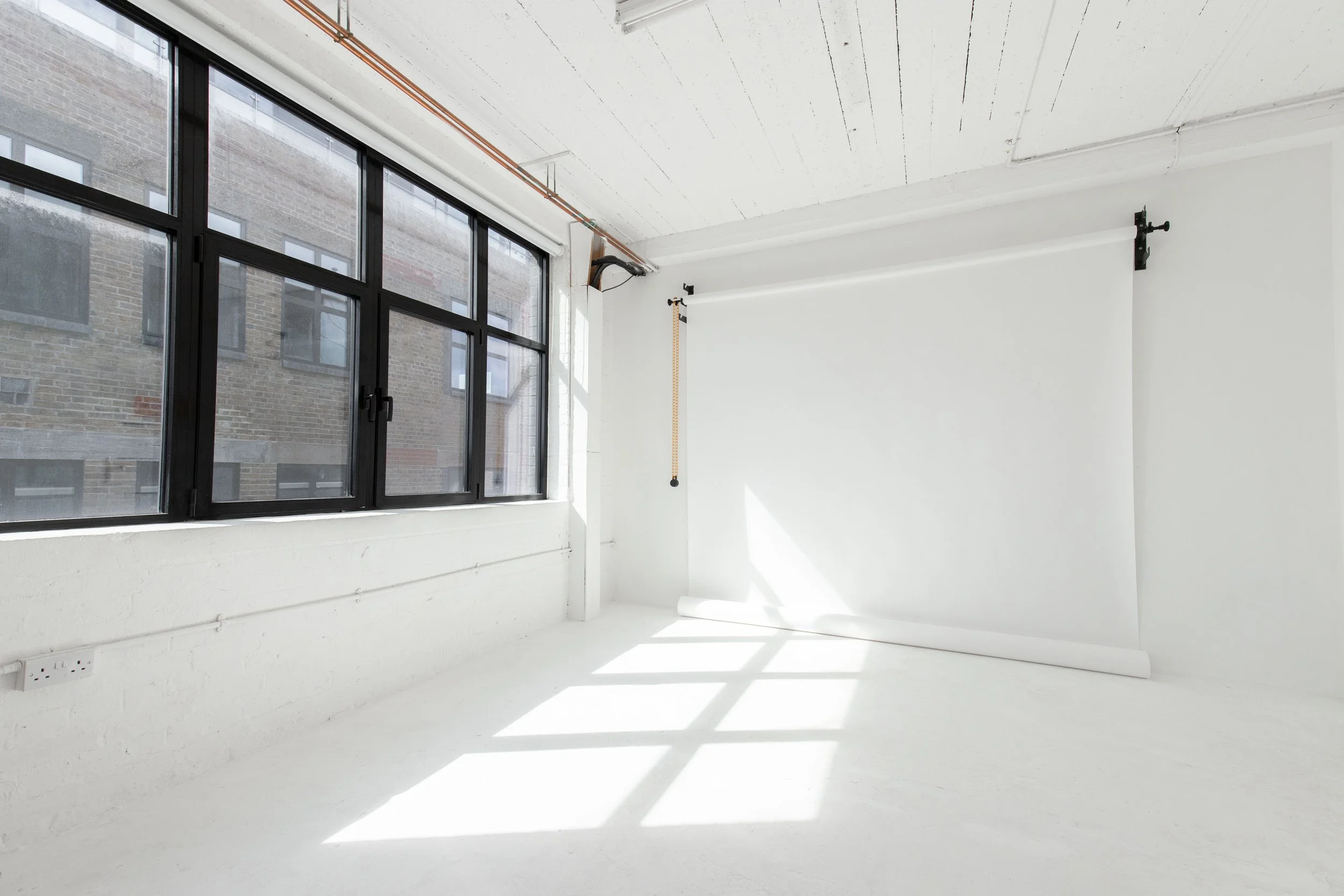 Empty photo studio with white walls, large black-framed window, natural sunlight, and a white backdrop on a stand.