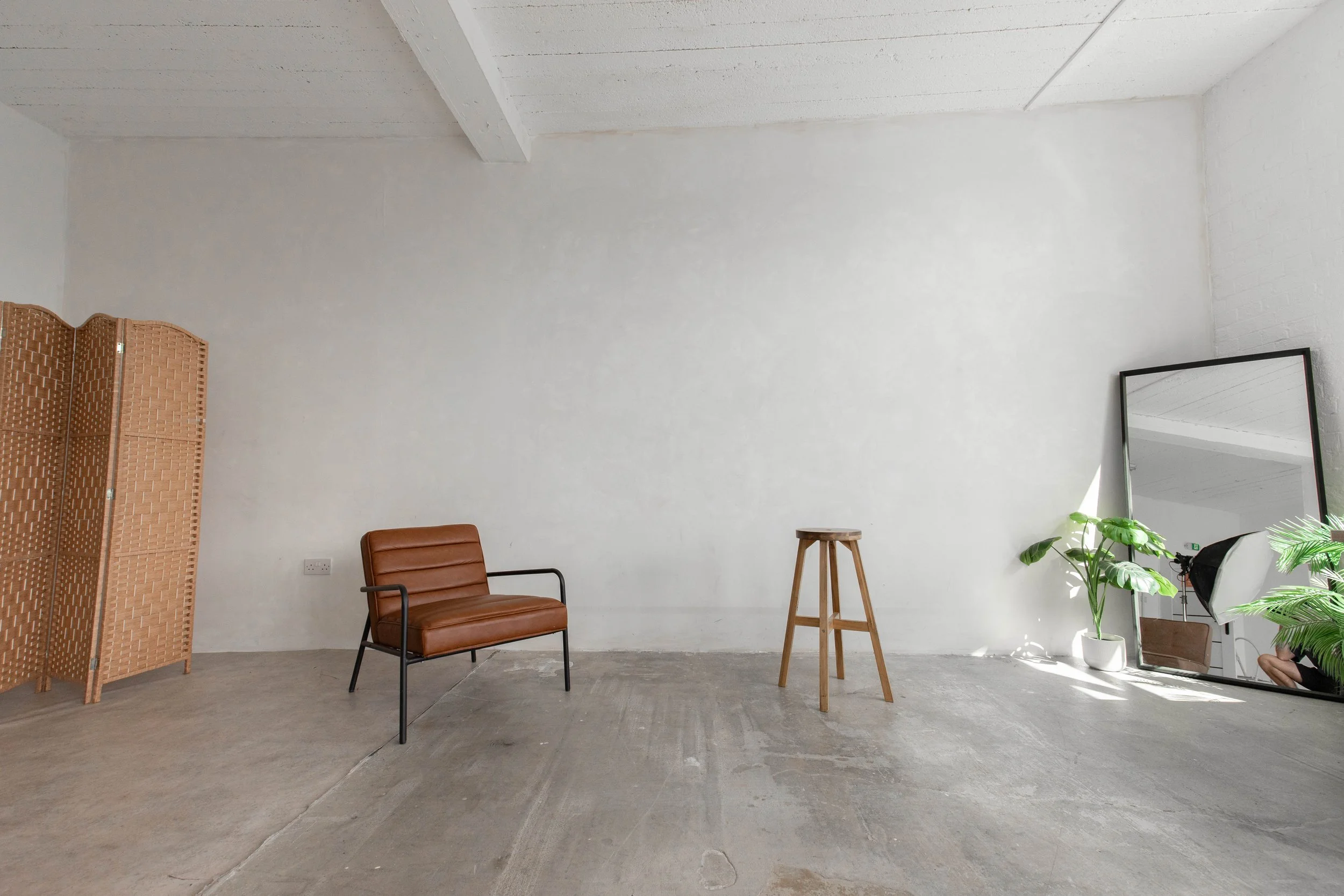 Minimalist room with a tan leather chair, a wooden stool, a large mirror leaning against the wall, and green plants in white pots.
